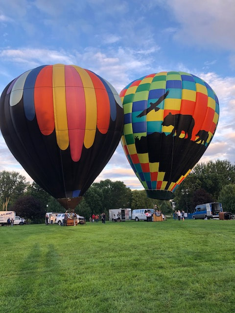 two hot air balloons tethered side by side, one with multi-color stripes over the top and the other with rainbow boxes and a silhouette of an eagle, a bear and a cub